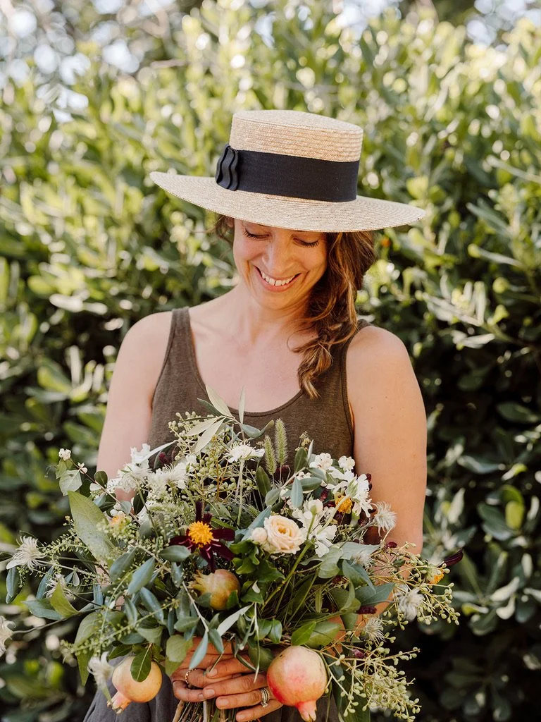 A woman in a brown sleeveless top wearing a straw hat with a black band, smiling while holding a bouquet of flowers and apples, outdoors with green foliage in the background.