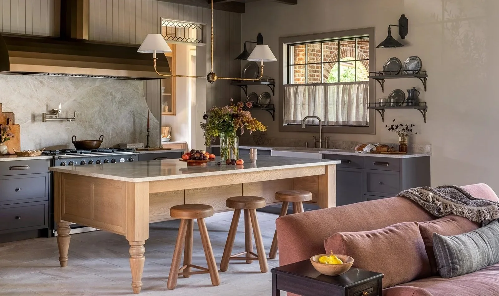 Cozy kitchen with a central island, wooden stools, and open shelving with dishes and cups. A window with curtains above the sink, a vase with flowers, and a couch with cushions nearby.