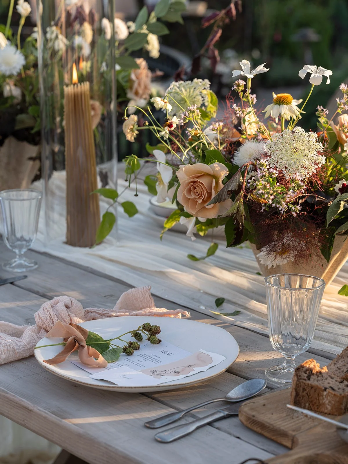 A rustic outdoor table set for a meal with a floral centerpiece, candles, glasses, and cutlery during sunset.