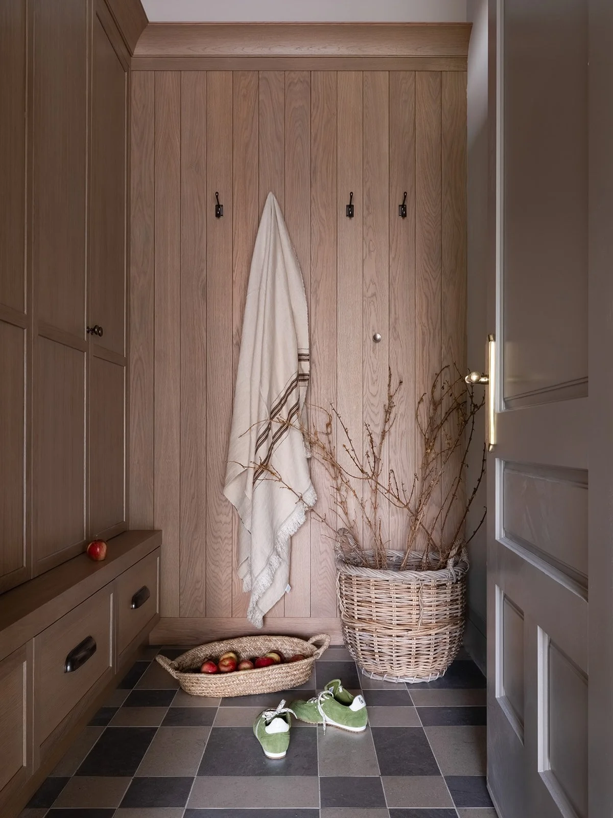 A cozy entryway with wood-paneled walls, a coat hook with a beige towel hanging, a large wicker basket with dried branches, a basket filled with apples, and a pair of green baby shoes on the checkered tile floor.