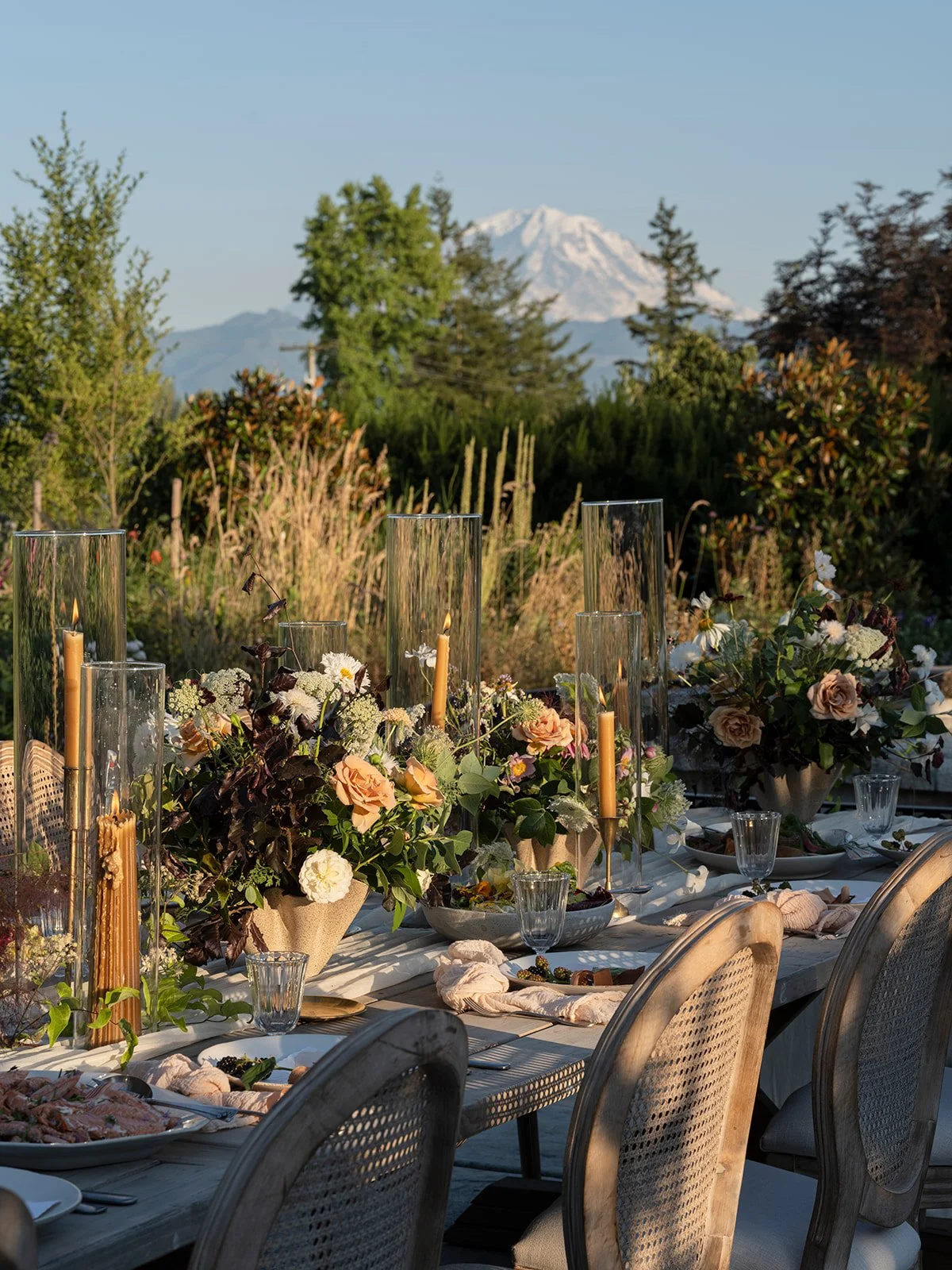Outdoor dining table set with floral arrangements and candles against a backdrop of trees and snow-capped mountain.