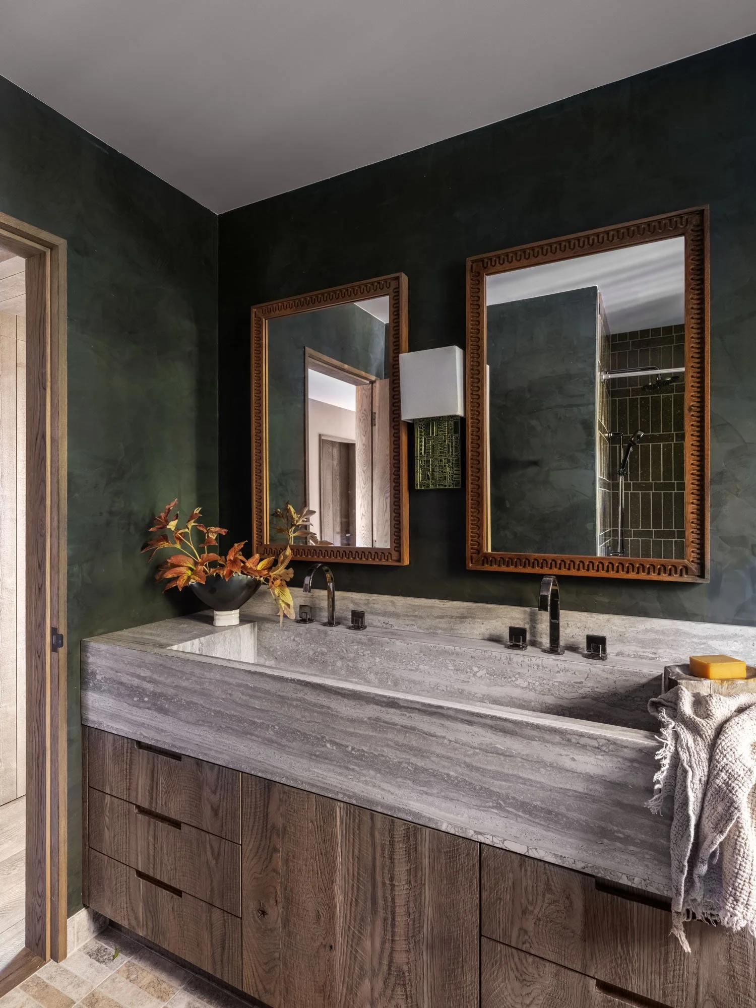 Double vanity with grey marble countertop, two rectangular mirrors with wooden frames, black fixtures, and a vase with autumn foliage, brown towels, soap, gray wall, and a reflection of a shower in the mirrors.