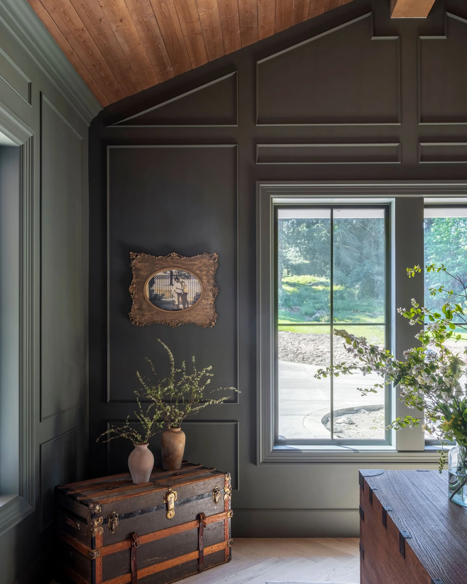 Interior corner of a room with dark green paneled walls, a large window, a vintage wooden trunk with leather straps and brass hardware, and decorative vases with greenery.