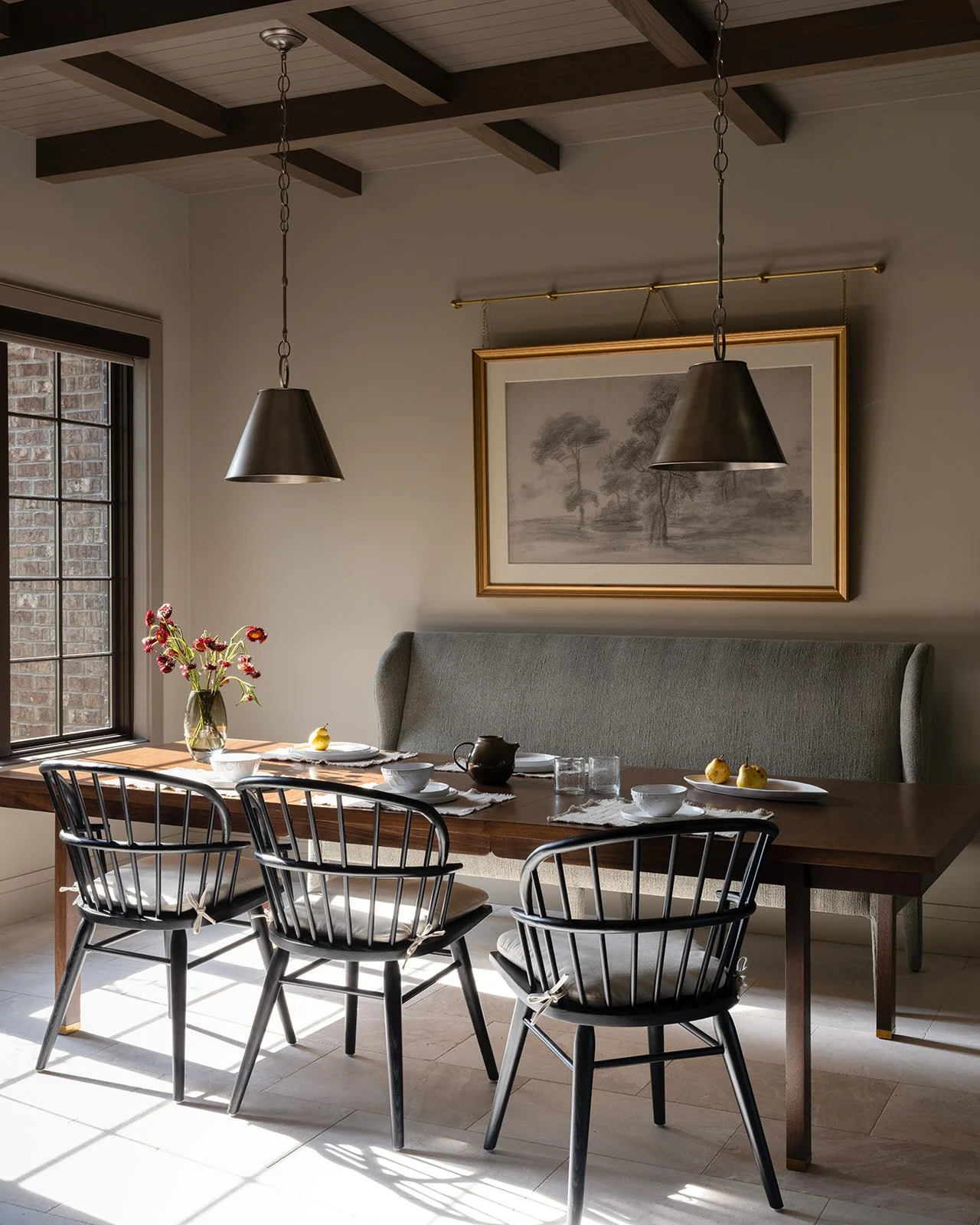 Dining room with a wooden table set for breakfast, black chairs with white cushions, a bench with a gray headboard, a vase with red and yellow flowers, and a landscape drawing on the wall.