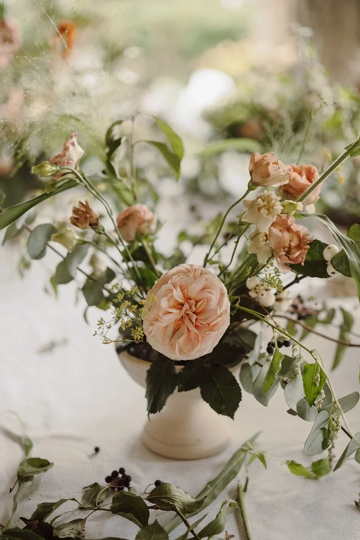A soft pink flower bouquet with greenery in a white bowl on a table, with scattered leaves and berries around it.