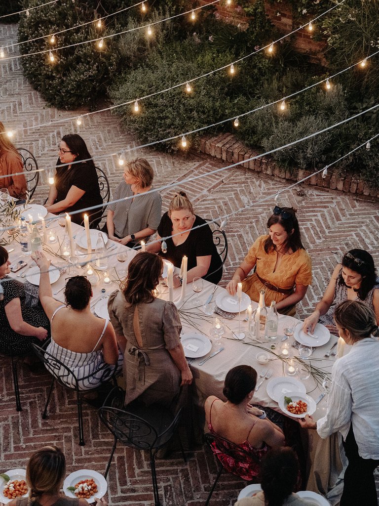 People sitting at a long dinner table outdoors, decorated with candles, string lights, and greenery, during an evening gathering.