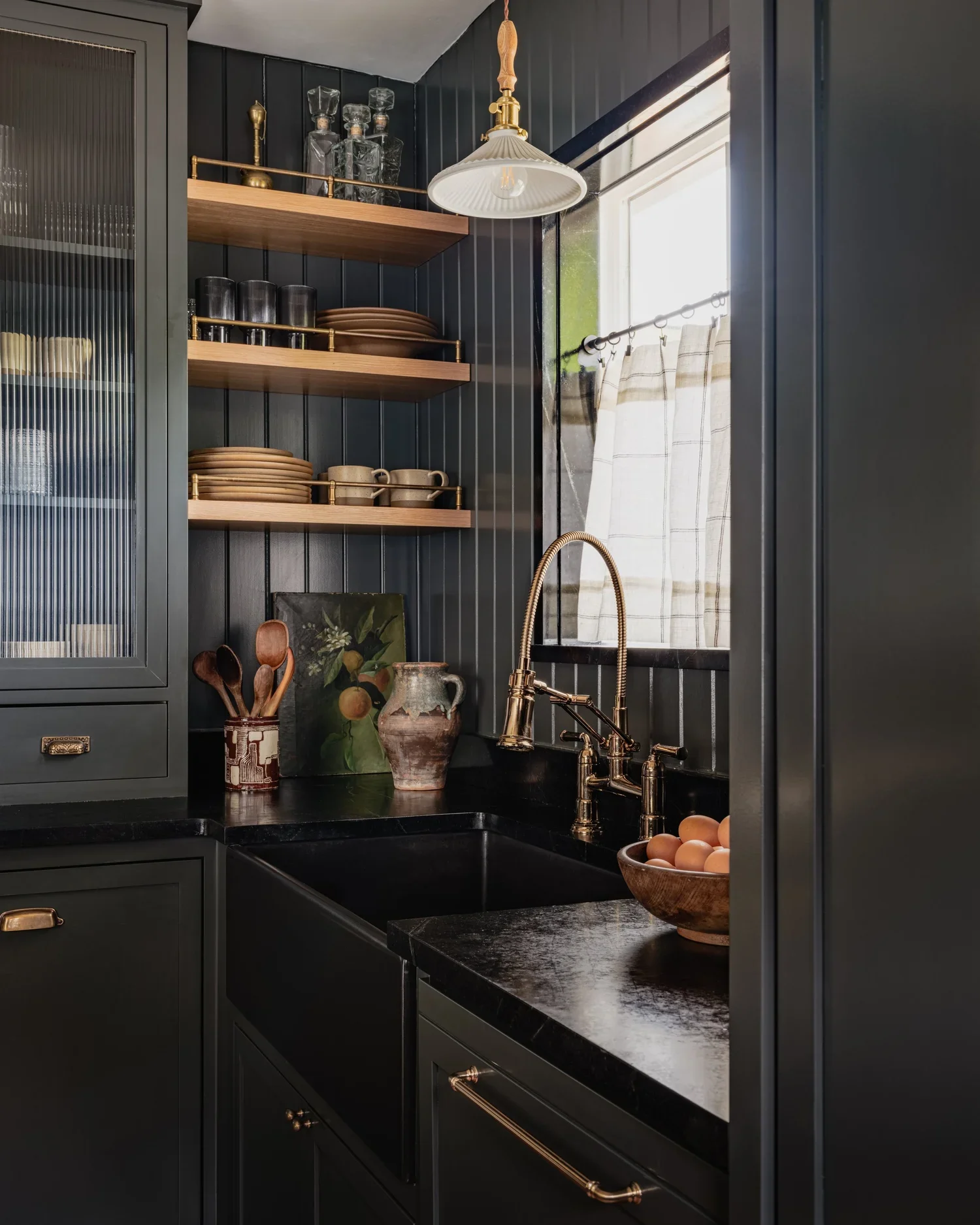 A cozy kitchen corner with dark green cabinets, open wooden shelves holding plates, cups, glasses, and decor, a black sink with a gold faucet, a bowl of eggs, and a window with a beige curtain.
