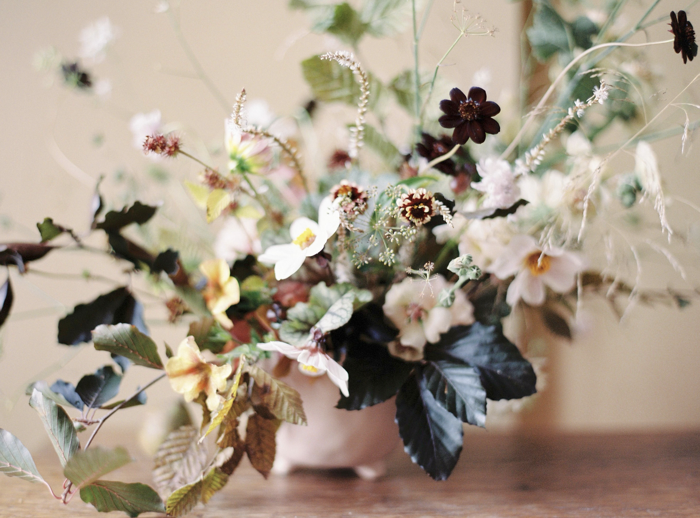 A bouquet of mixed flowers in a bowl on a wooden surface, including white, pink, yellow, and dark purple blooms with green and dark leaves.