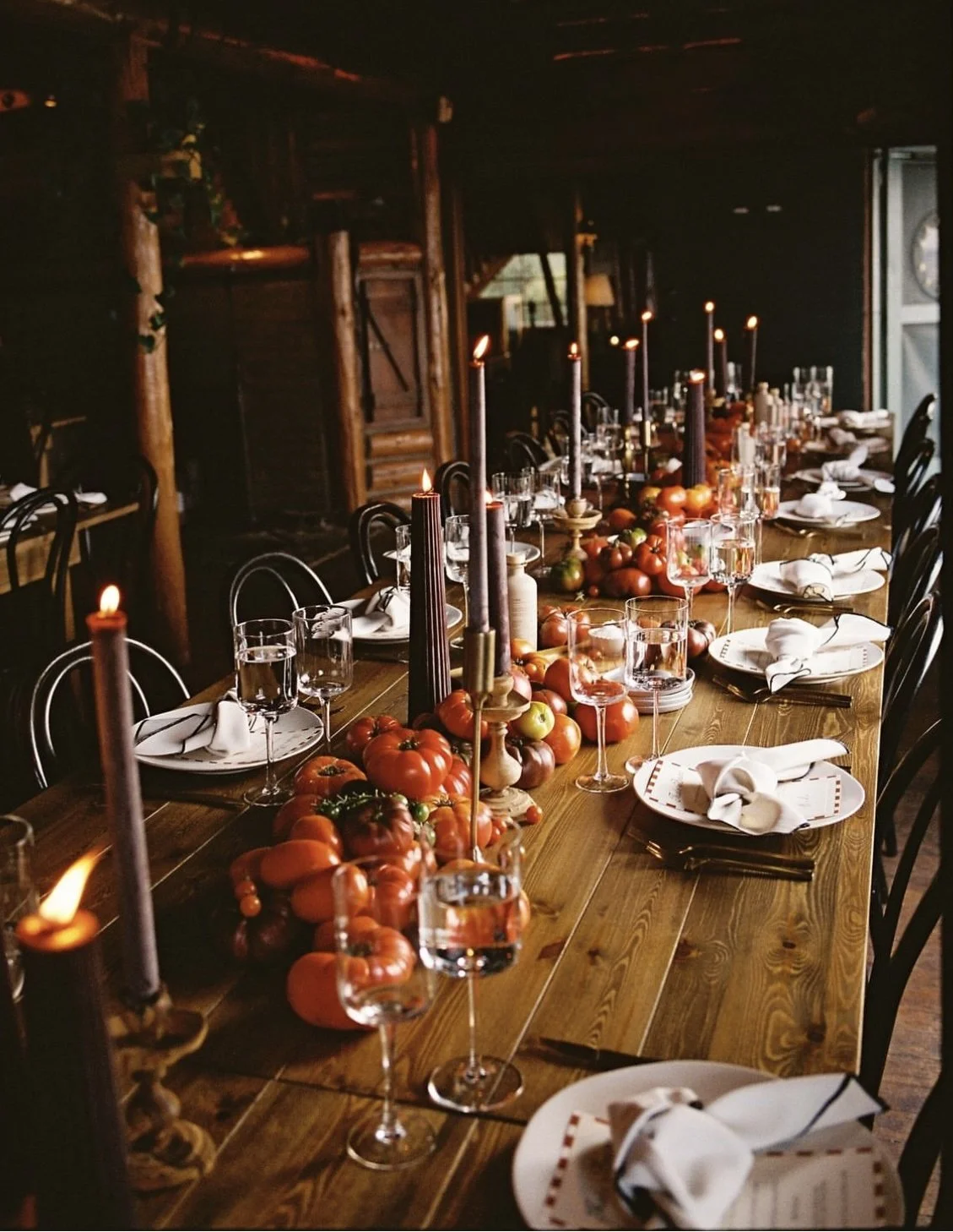 A long wooden dining table set with white plates, cloth napkins, and empty wine glasses, decorated with bunches of tomatoes and lit candles in a rustic, cozy room.