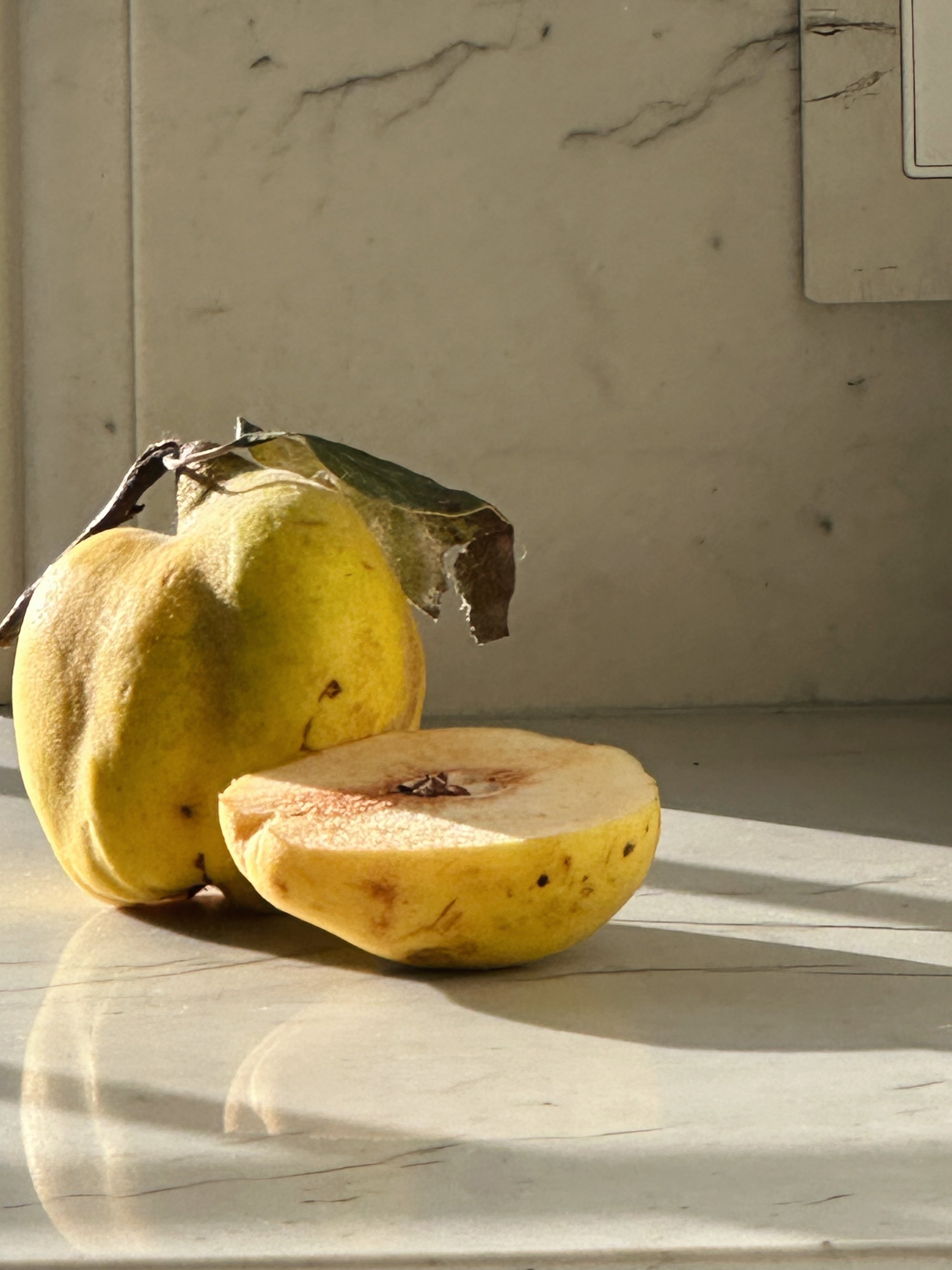 A yellow apple, one half-cut and one whole, with a dry leaf attached, sitting on a light-colored surface with shadow, near a wall with visible lines and a metallic object in the background.