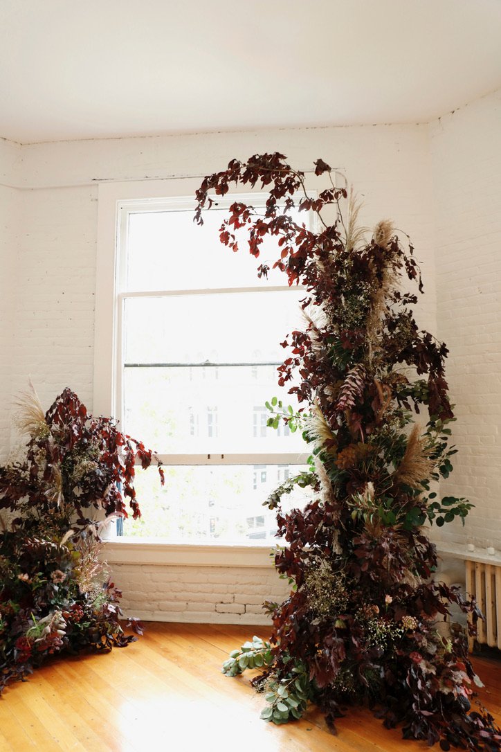Indoor scene with large window and tall floral arrangements made of dark red and green foliage, dried grasses, and small white flowers.