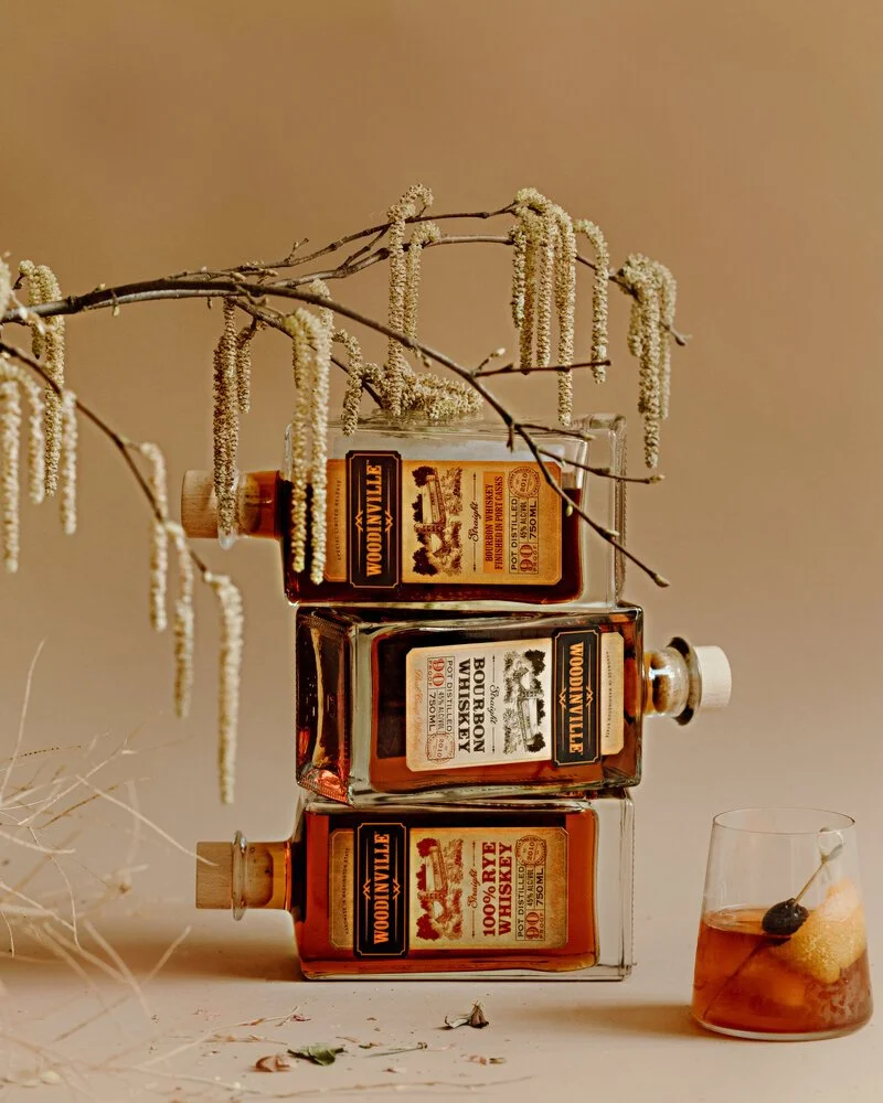 Three bottles of Woodinville bourbon whiskey stacked and a glass of whiskey with ice and garnish on a table, with dry branches and flowers as decoration.