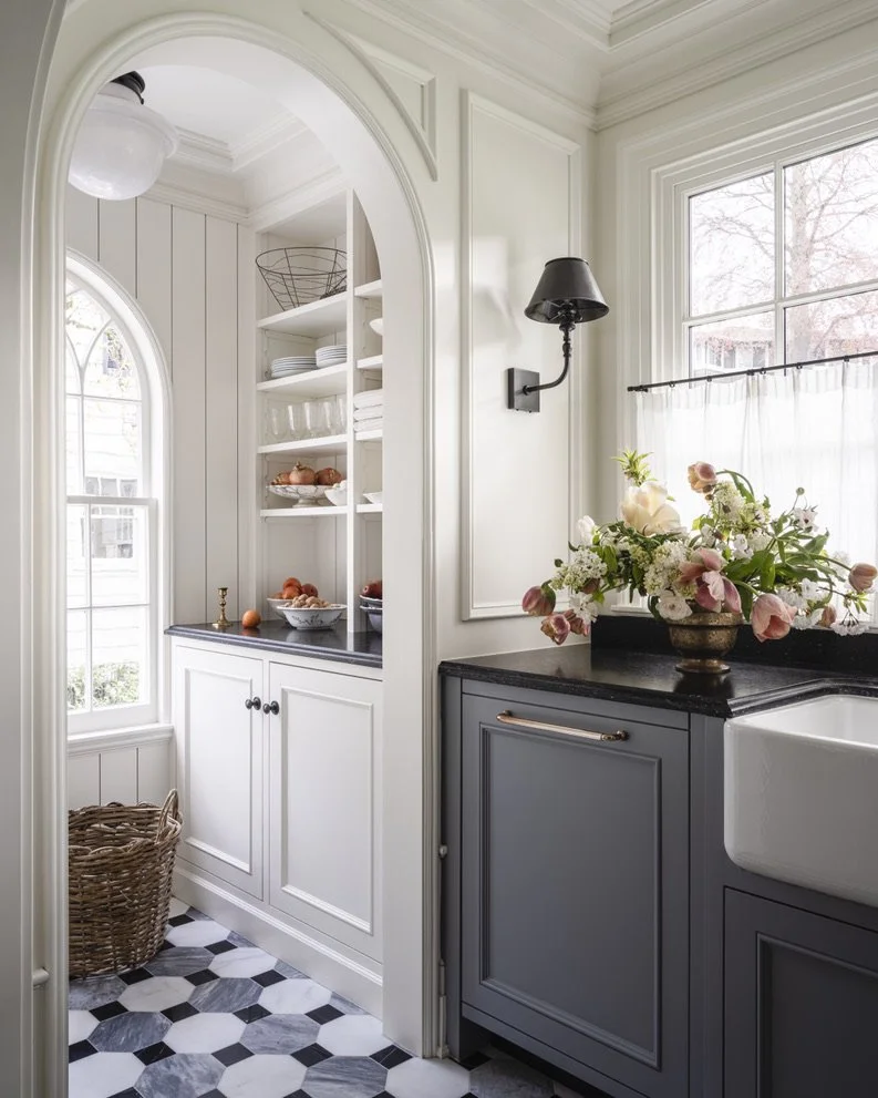 A cozy kitchen corner with white cabinets, a black countertop, and a gray lower cabinet next to a farmhouse sink. There is a window with sheer curtains, a bouquet of flowers on the counter, and open shelves with dishes and bowls.