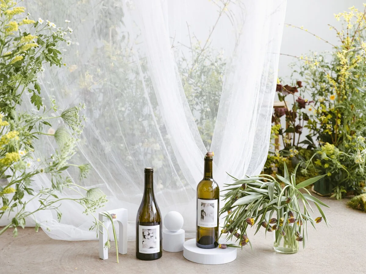 Empty wine bottles, white decorative objects, and a vase with plants on a table in a bright room with curtains and yellow and green plants in the background.
