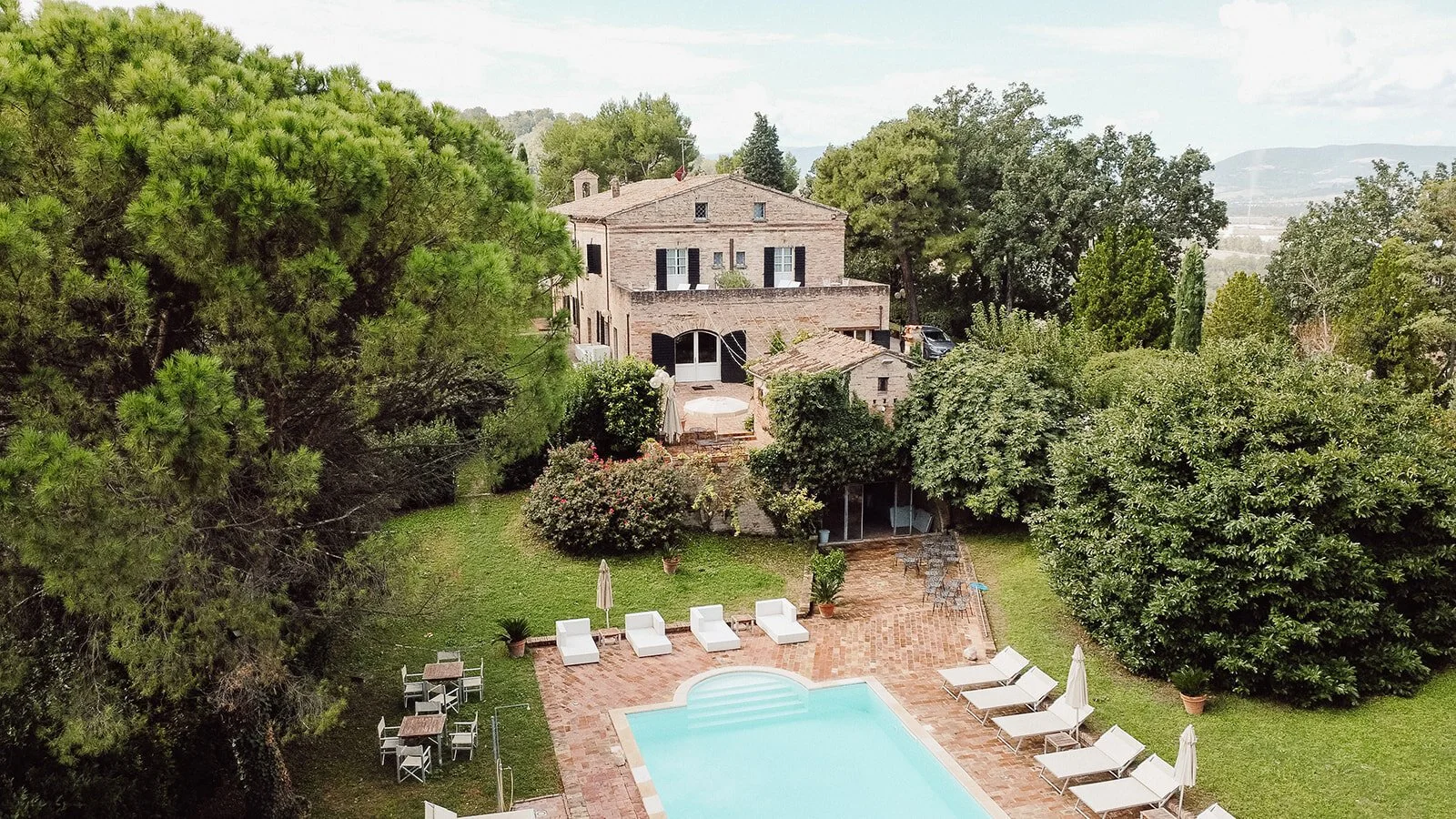 Aerial view of a backyard featuring a swimming pool with lounge chairs, side tables, umbrellas, lush green trees, a stone patio, and a two-story brick house in the background.