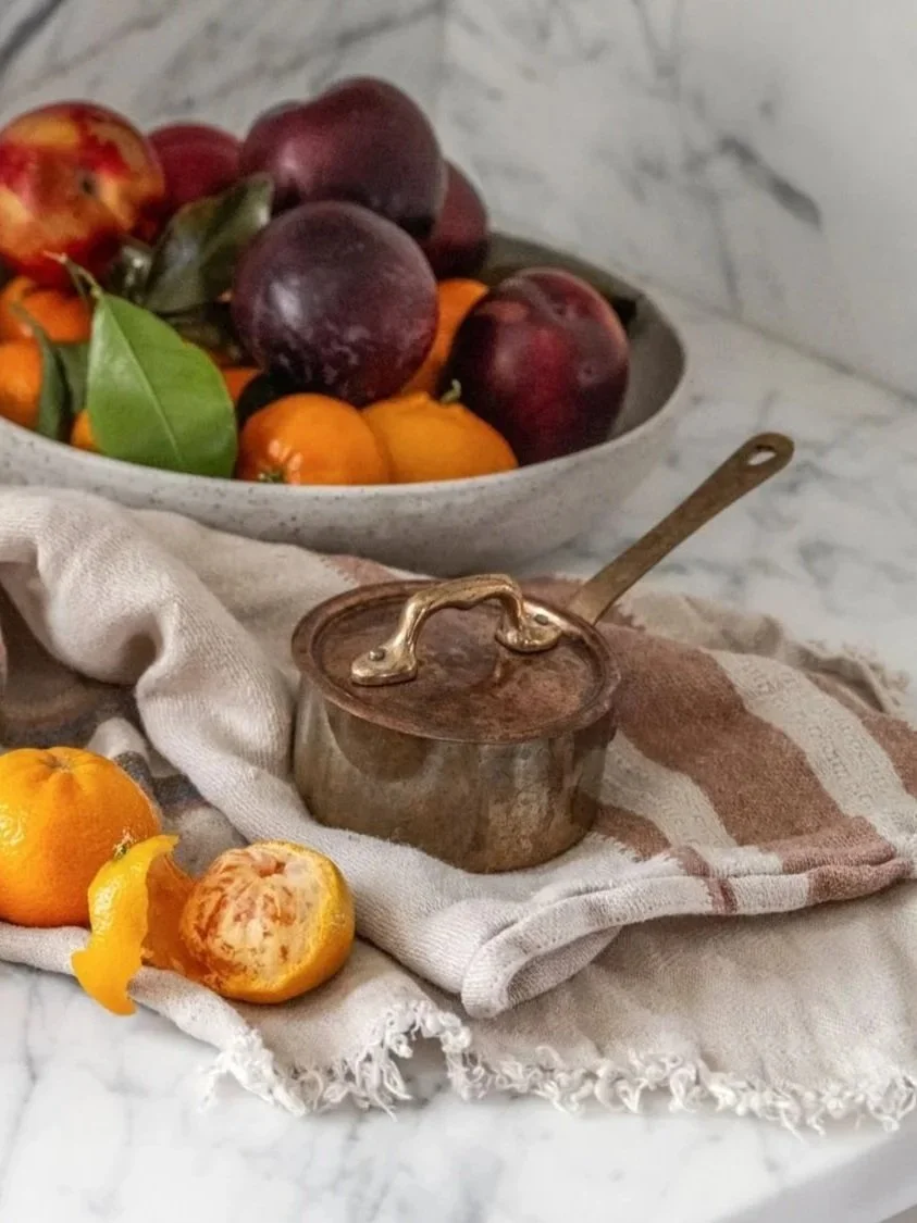 A bowl of assorted fresh fruit, including peaches, nectarines, and oranges, with a partially peeled orange in the foreground, set on a marble surface with a rustic cloth underneath.