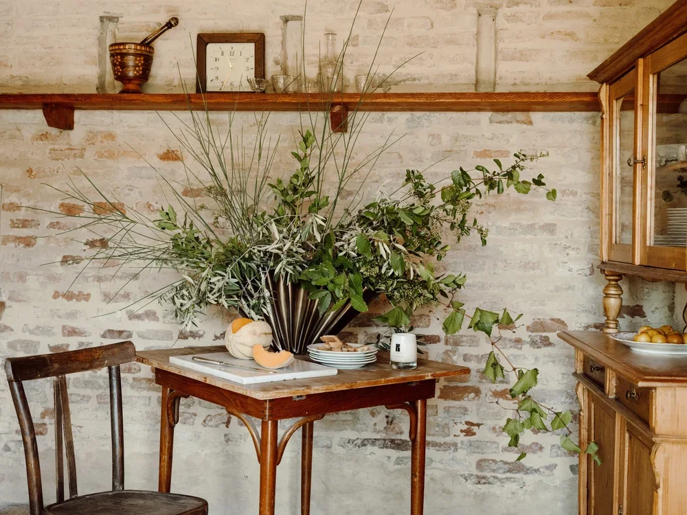 A wooden table with green plants in a large vase, a melon, plates, and a candle in front of a textured brick wall in a rustic kitchen or dining area.