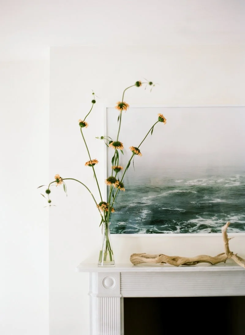 Vase with tall flowers and a piece of driftwood on a white mantel, with a framed ocean photograph behind.