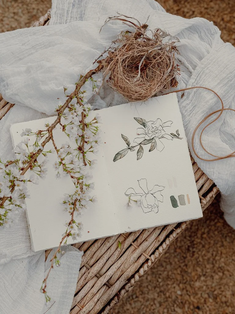 A sketchbook with floral sketches and color swatches, a branch with white blossoms, a dried plant, and light-colored fabric on a woven surface.