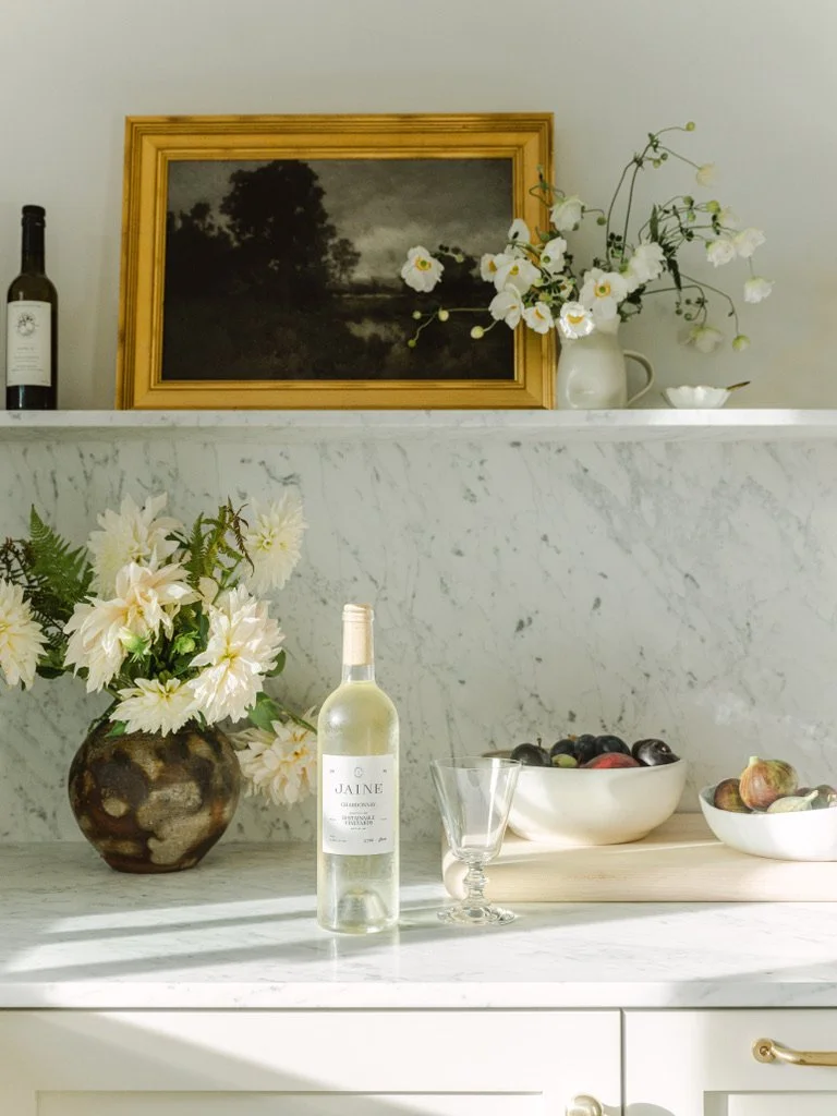Decorative kitchen shelf with a framed landscape painting, white flowers in a pitcher, a vase with white flowers, a bottle of white wine, two bowls of fruit, a glass, and a small bowl on a white marble surface.