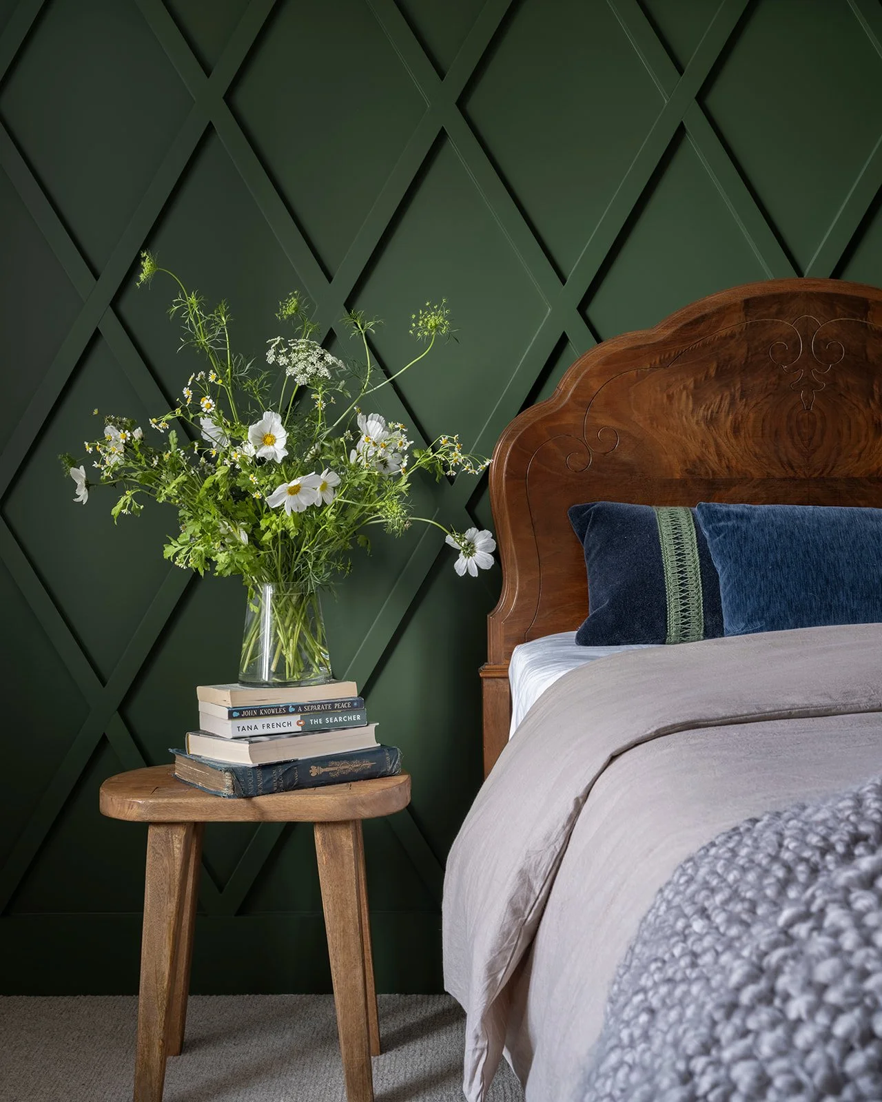 Bedroom with a wooden headboard, green wall with a diagonal pattern, a bedside table with books and a glass vase of white and green flowers.