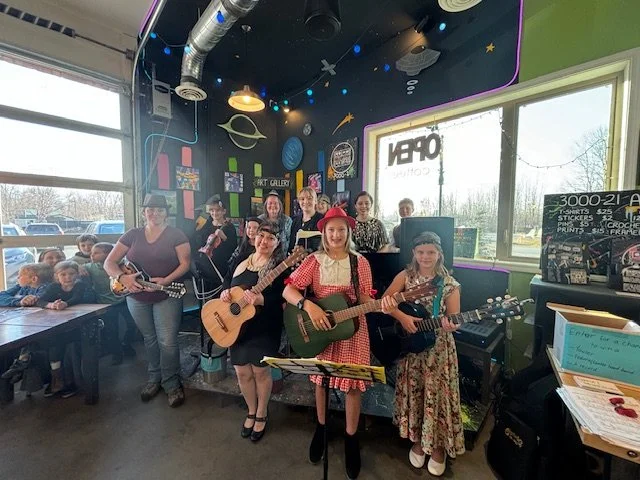 Five women, some with guitars, performing in a colorful, space-themed mural cafe, while children sit at a table nearby.