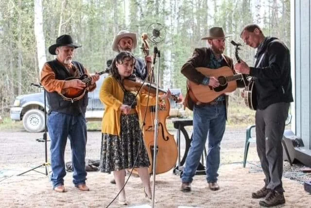 Group of five people, four men and one young girl, playing musical instruments outdoors, with a backdrop of trees and a parked car.