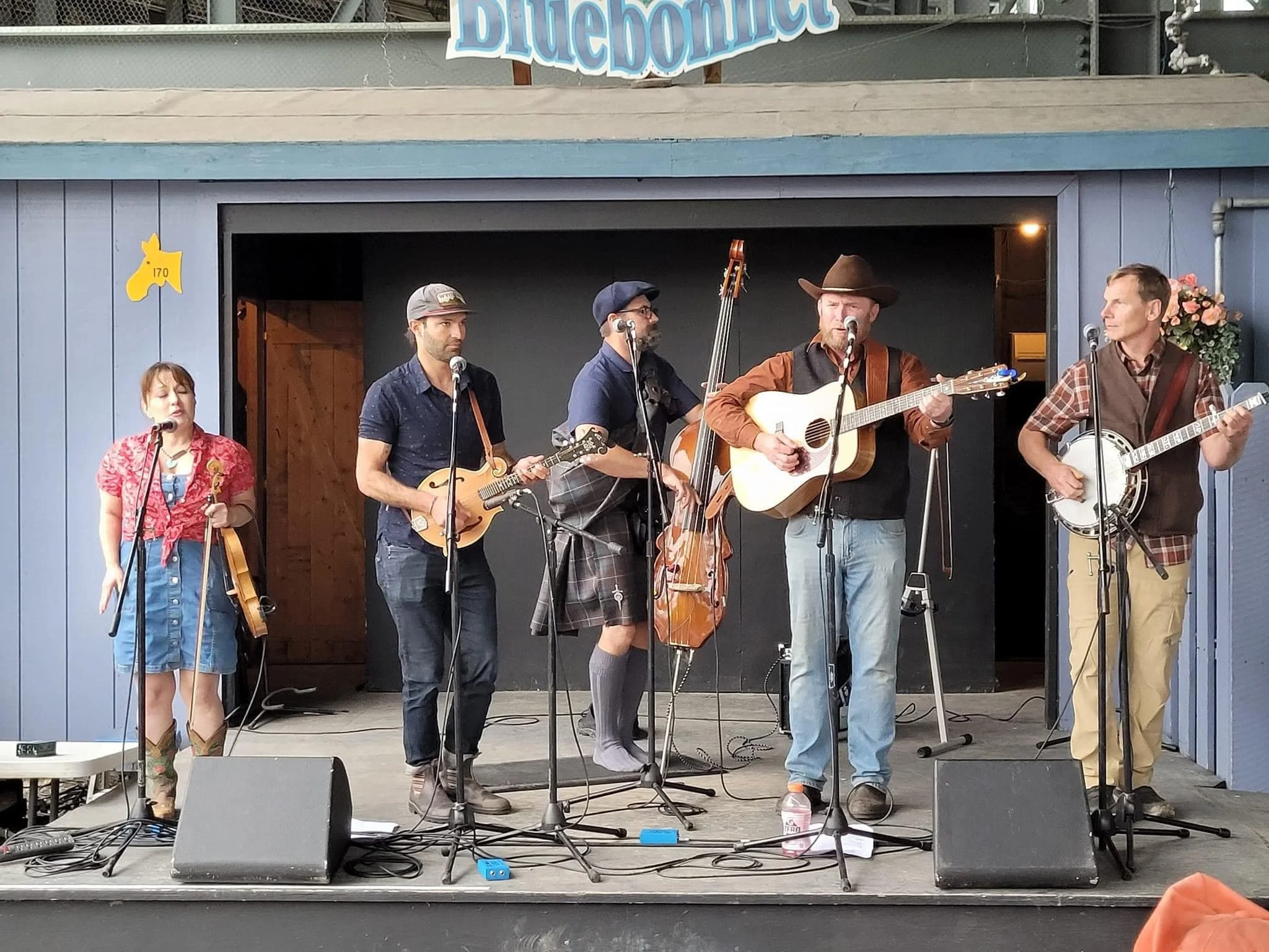 A band performing on an outdoor stage, includes five members with various instruments, under a sign that reads 'Bluebonnet'.