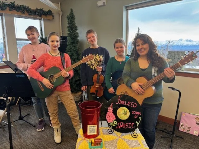 Five children and a woman with guitars in a classroom, with a sign that reads "Fireweed School of Music."