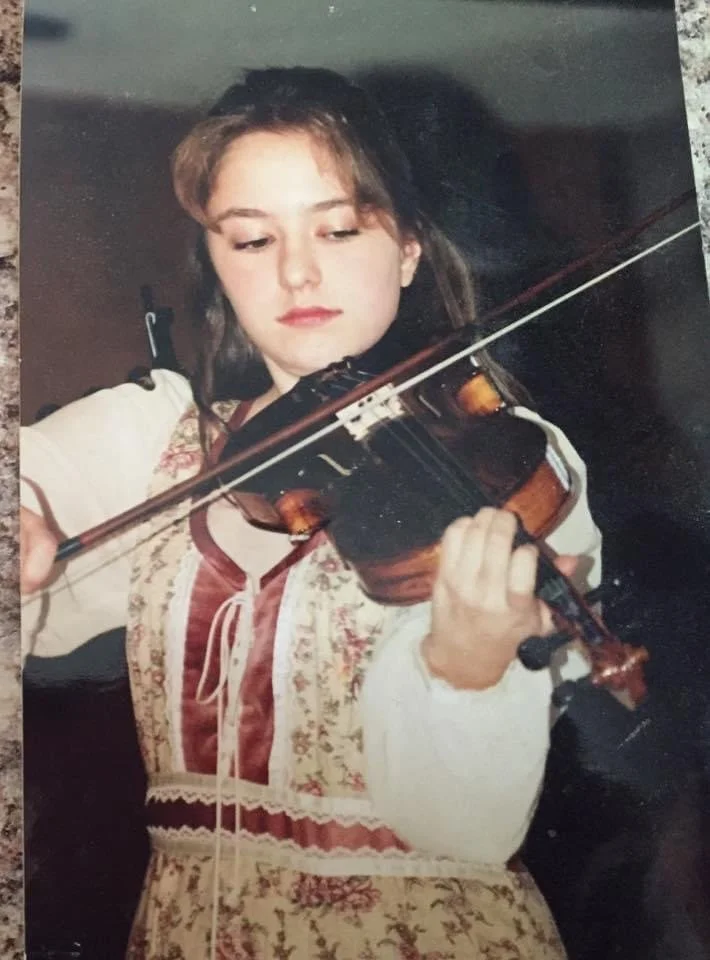A young girl with brown hair playing a violin. She is wearing a traditional embroidered dress.