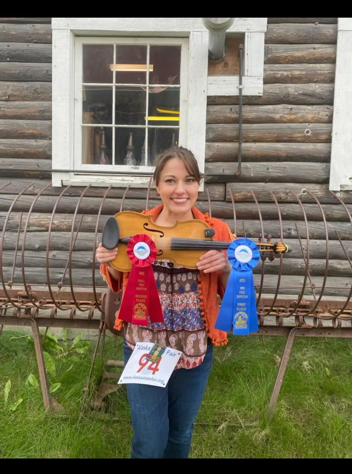 A woman holding a violin with red and blue award ribbons, standing outdoors in front of a rustic wooden building, with a grass lawn at her feet.