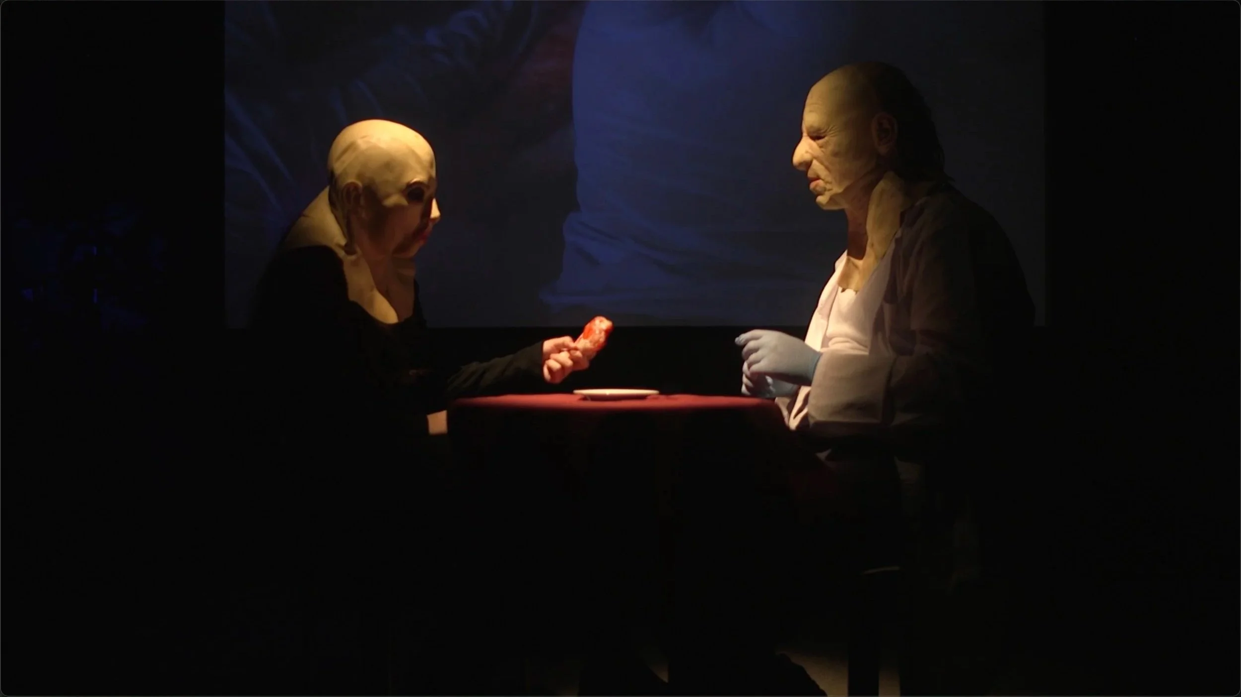 Two people wearing masks with exaggerated facial features sit at a small table in a dimly lit room, with one holding a piece of food and the other wearing gloves, possibly in a theatrical or artistic setting.