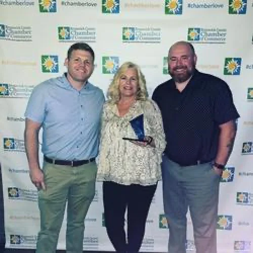 Three people standing together, smiling in front of a backdrop with logos and text.