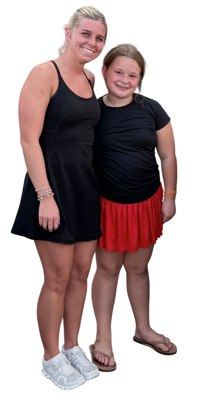 Two smiling women stand close together, wearing casual summer outfits. One has blonde hair tied back, wears a black tank dress and sneakers. The other has brown hair, wears a black t-shirt, red skirt, and flip-flops.