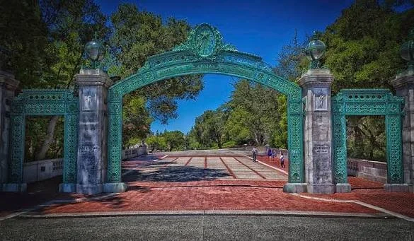 Entrance with an ornate green archway and stone pillars, leading into a park with trees and pathways under a clear blue sky.