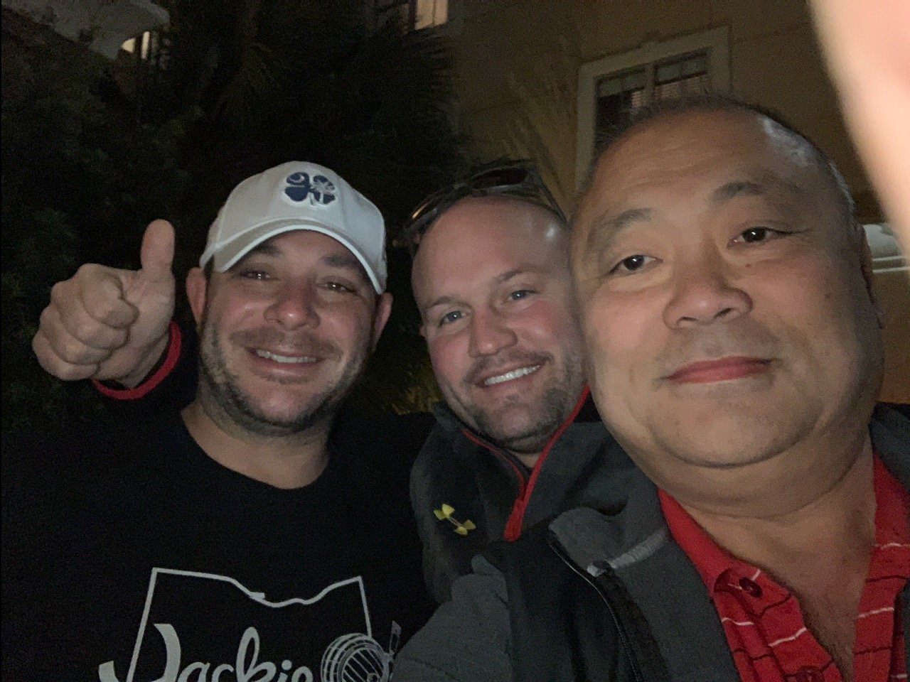 Three men smiling and taking a selfie together at night. One of them is giving a thumbs-up. They are outdoors or in a semi-outdoor area.