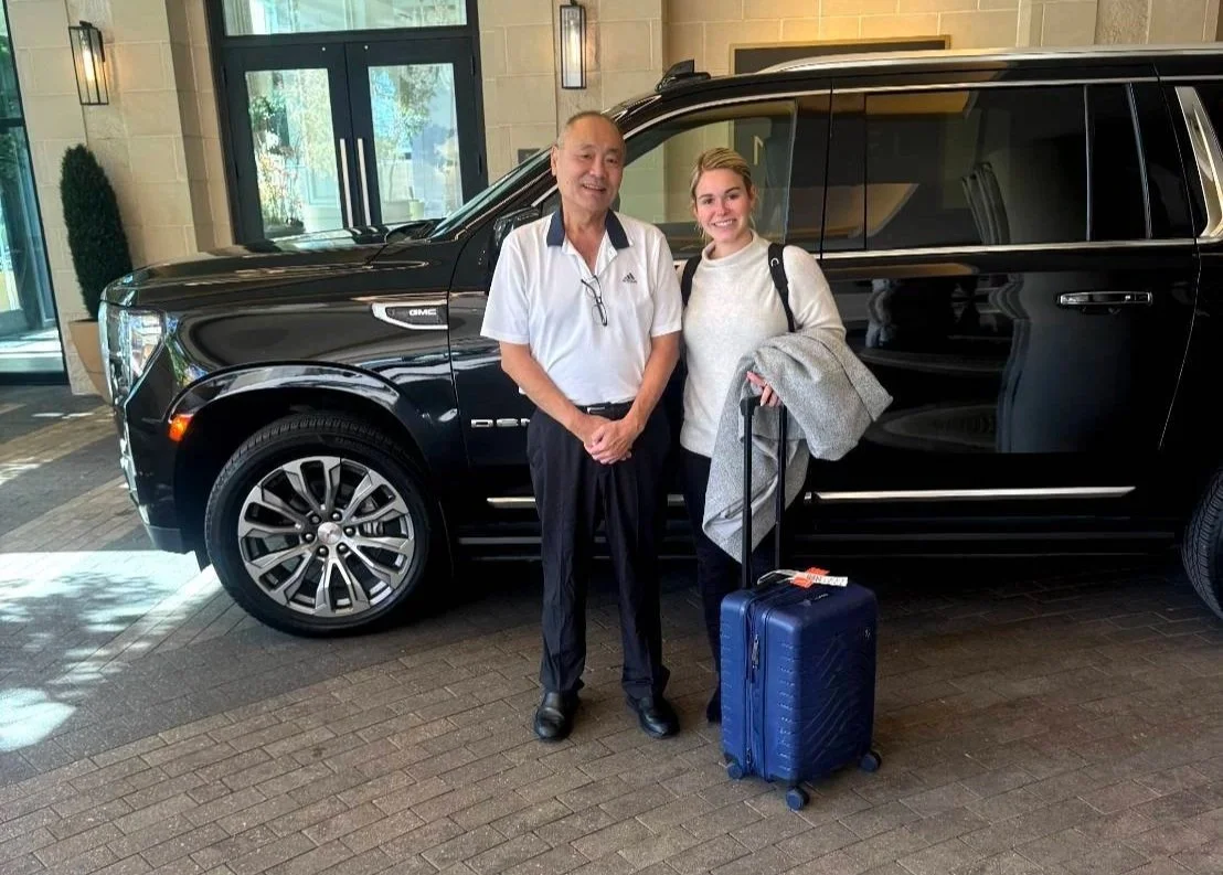 A man and a woman standing in front of a black SUV in a hotel lobby, with the woman holding a rolling suitcase.