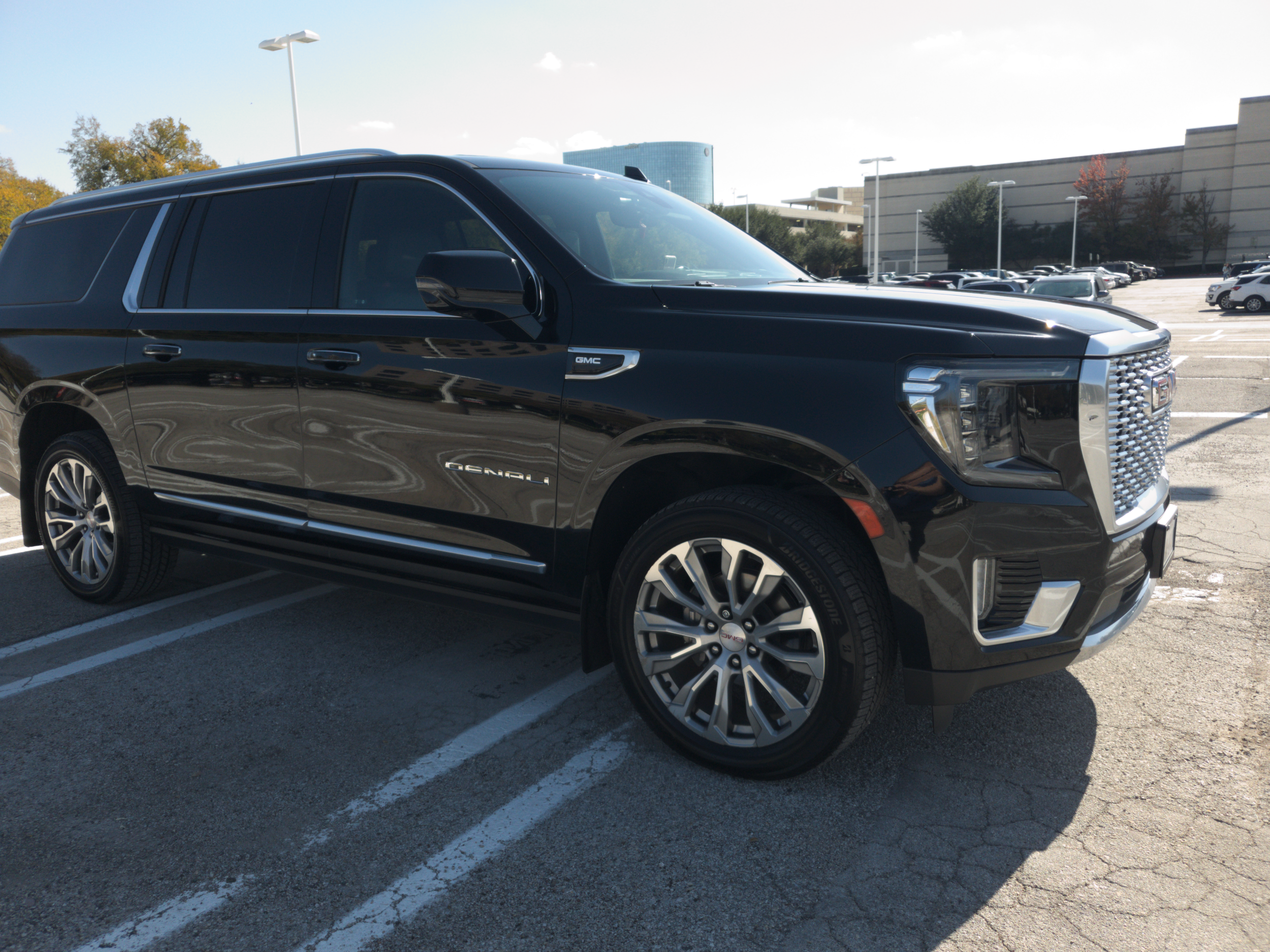 Black GMC Yukon Denali SUV parked in a parking lot on a sunny day, with other cars and a large building in the background.