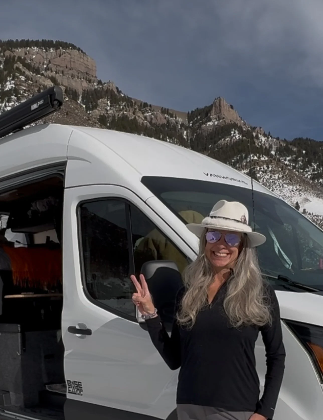 Woman with long gray hair, wearing a white hat, sunglasses, and black long-sleeve shirt, making a peace sign in front of a white van in a mountainous area with snow and trees.