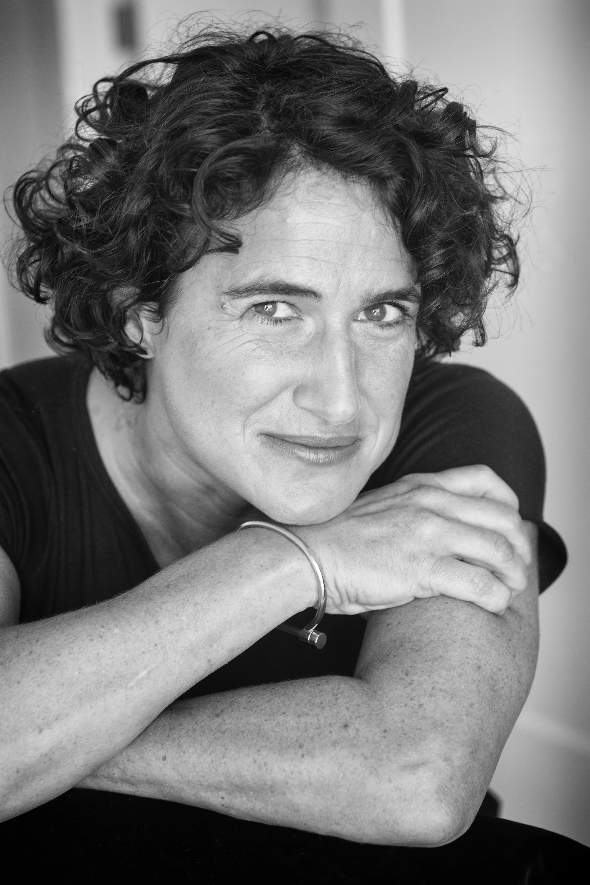 Black and white portrait of a woman with curly hair, resting her chin on her crossed arms, looking at the camera with a slight smile.