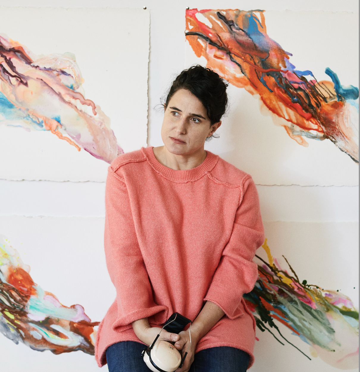 A woman with short black hair wearing a pink sweater sits in front of abstract colorful paintings on a white wall, holding a vintage camera.