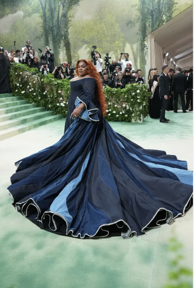 A woman in a flowing dark blue gown with light blue accents walking on a red carpet at a formal event, with photographers and attendees in the background.