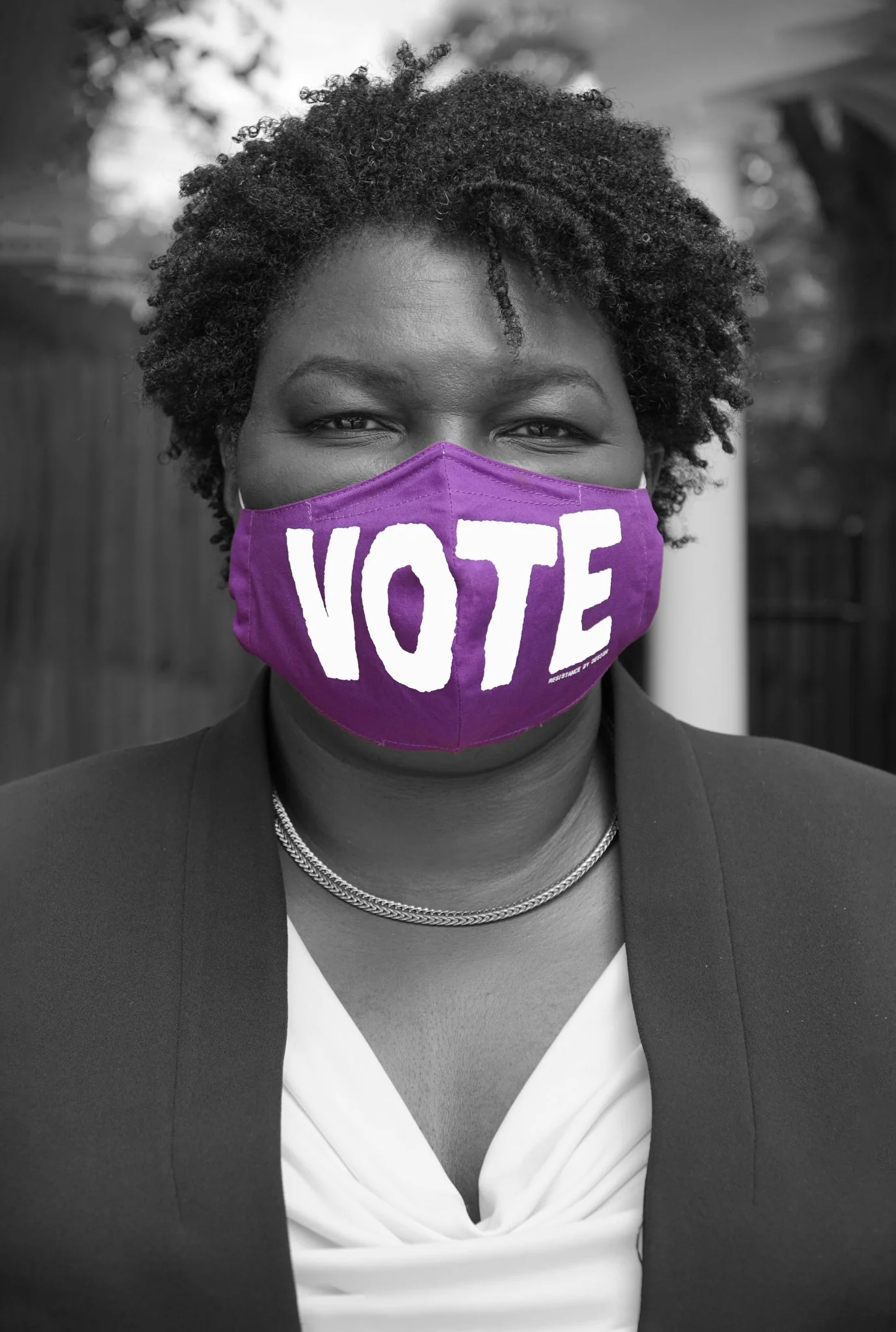 A woman with short, curly hair wearing a purple face mask with the word 'VOTE' written on it in bold white letters. The photo is black and white except for the mask.