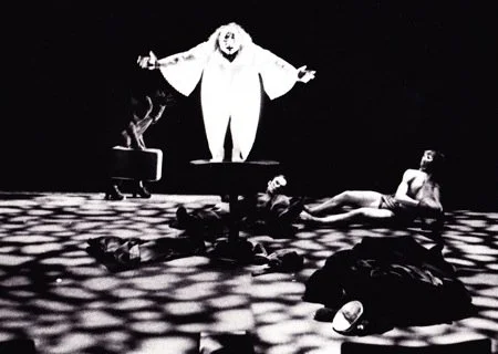 A stage scene with a person dressed as a clown or mime standing with arms outstretched, four people lying on the floor, and a person sitting near a cat on a raised platform.
