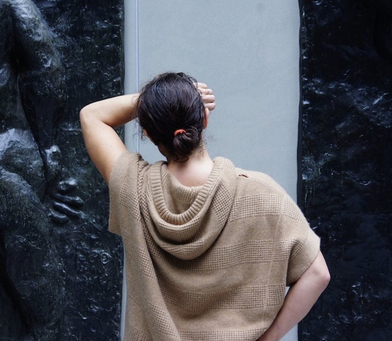 Back of a woman with dark hair tied in a bun, wearing a brown hooded sweater, standing with one arm behind her head, facing a gray panel and dark textured rocks.