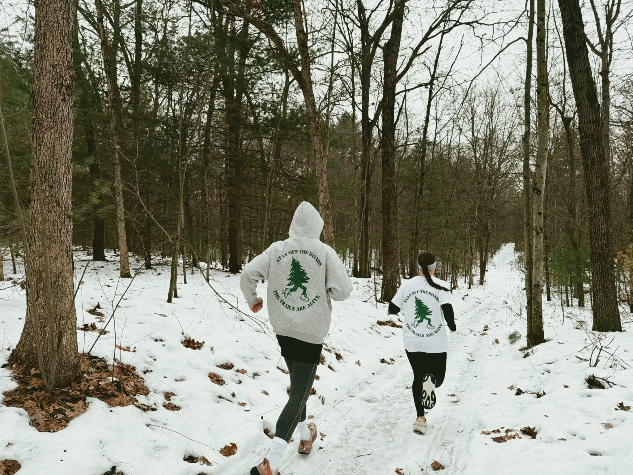 Two people running on a snowy forest trail, wearing matching gray sweatshirts with green trees and the phrase 'Stay off the roads, the trails are alive.'