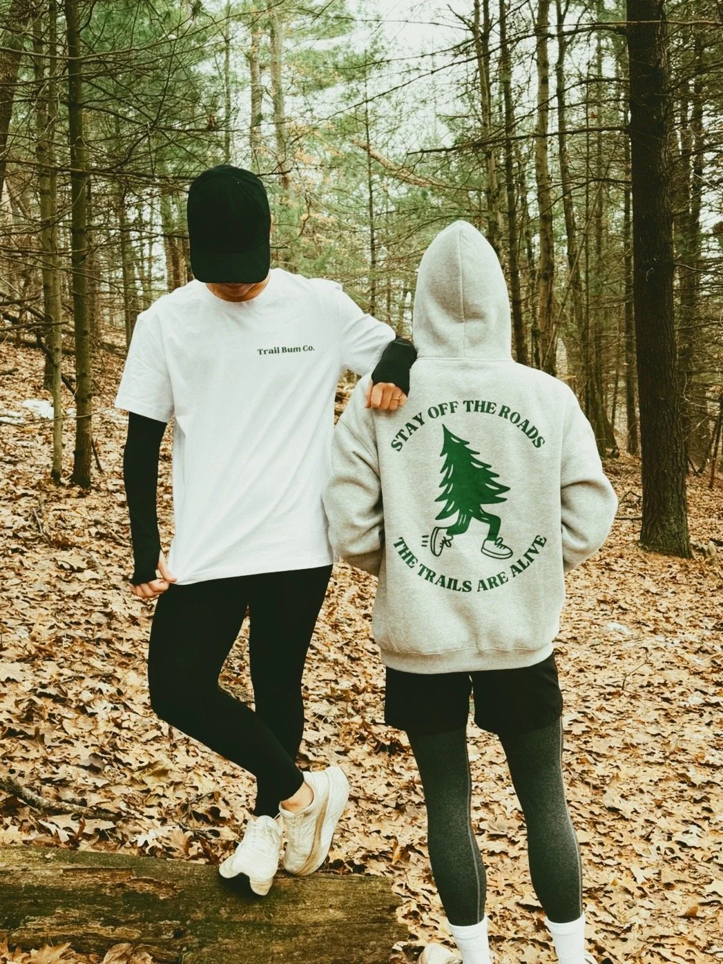 Two people standing on a trail in a wooded forest, with leaves on the ground. One person is wearing a white t-shirt, black cap, and black leggings. The other is wearing a gray hoodie with a graphic of a walking tree and the words "Stay Off The Roads, The Trails Are Alive" on the back.