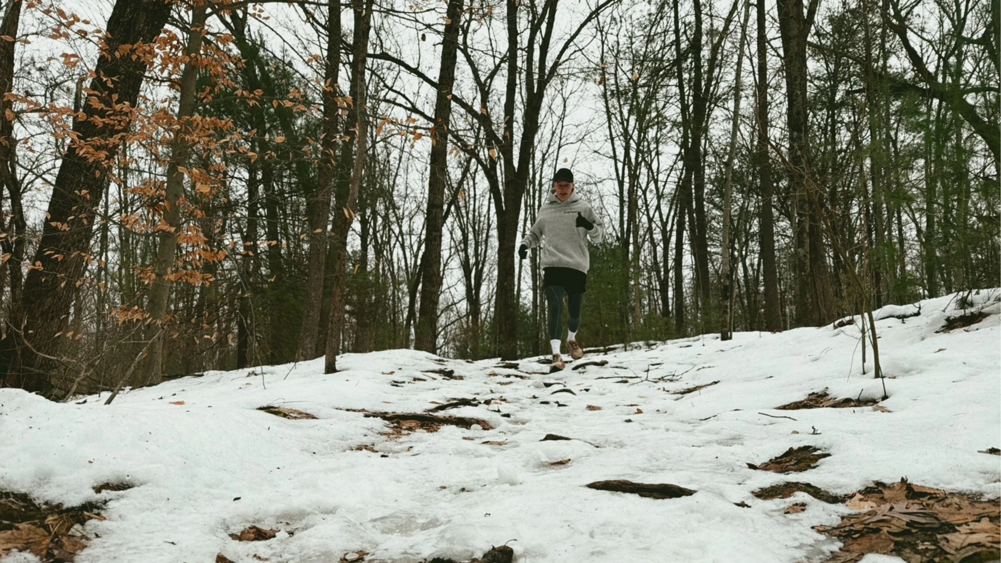 A person running on a snowy trail in a wooded area, wearing a light gray hoodie, black leggings, gloves, and a cap.