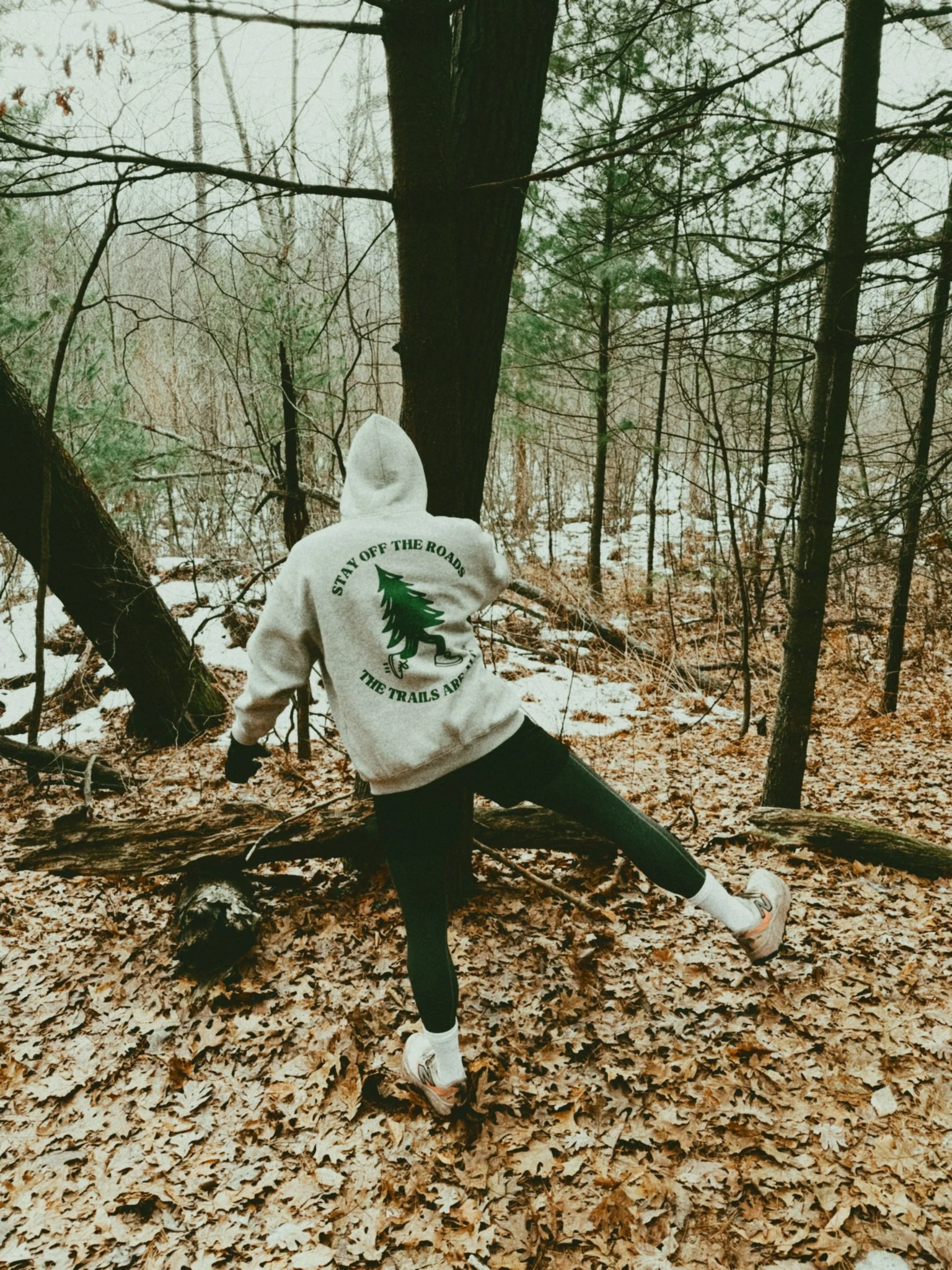 Person wearing a gray hoodie with a green pine tree logo and the words 'Stay Off The Roads, The Trails Are Free' hiking in a forest during winter.