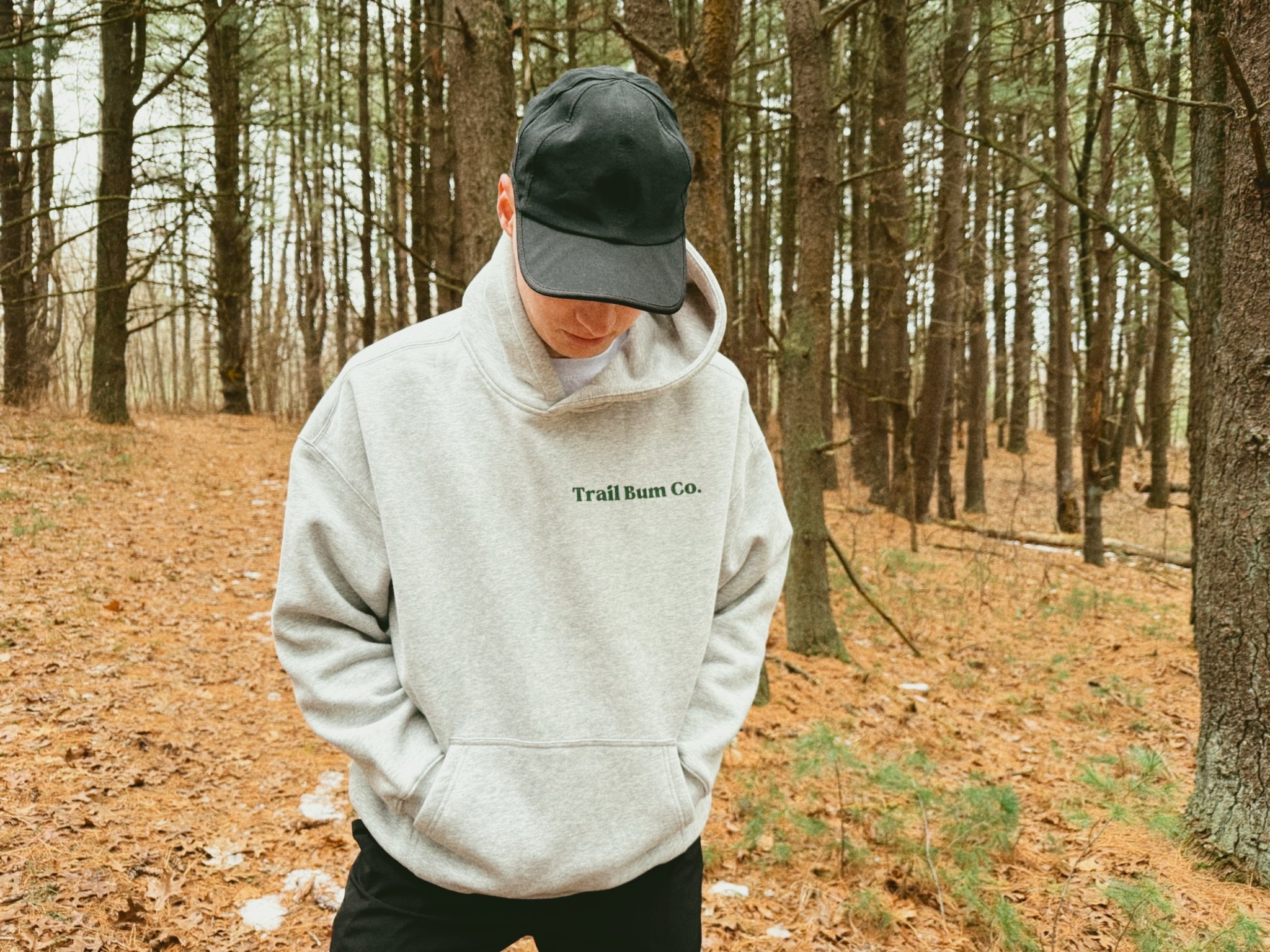 Person in gray hoodie with green text and black cap walking in a forest with orange fallen leaves.